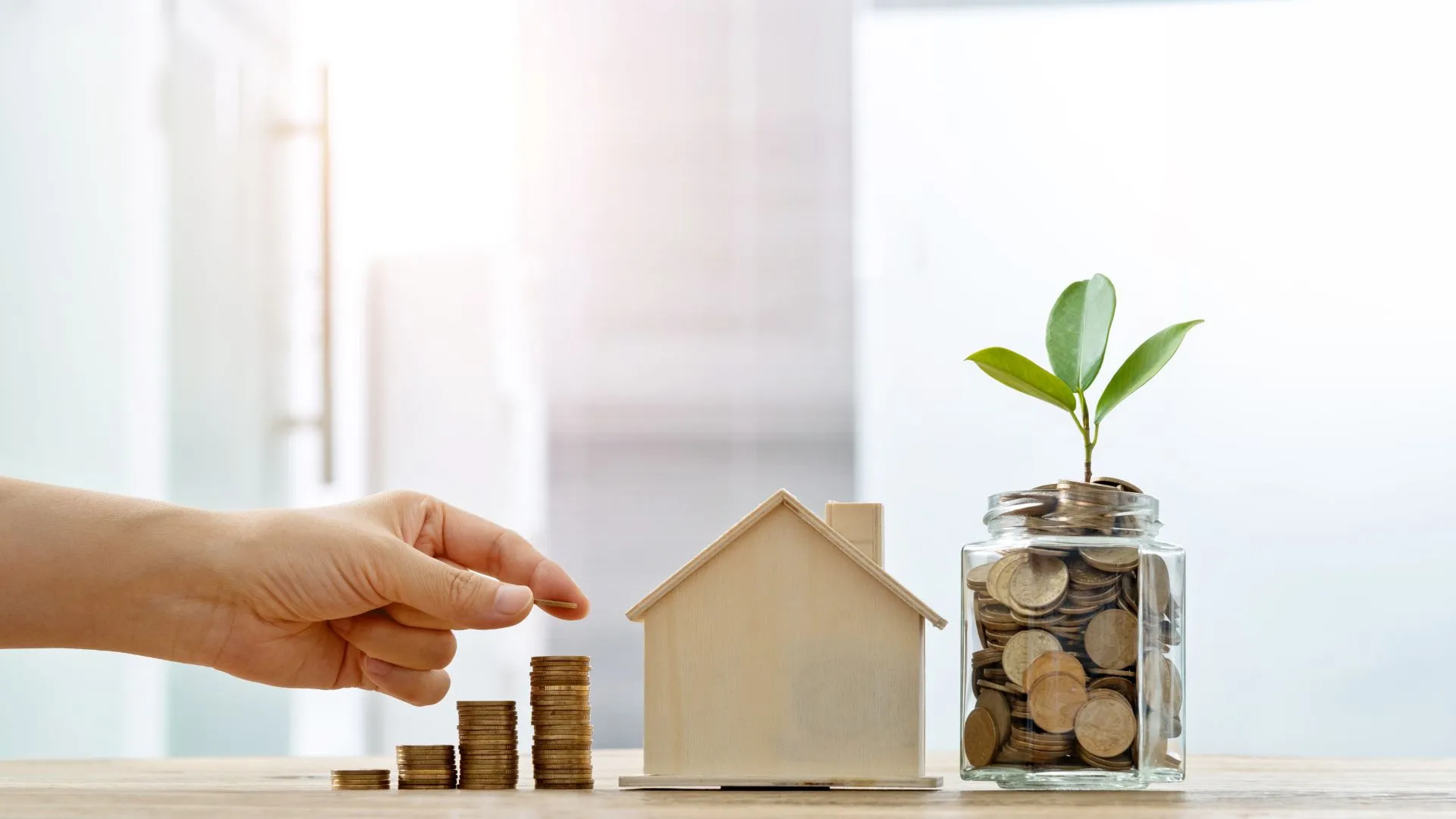 Hand stapelt Münzen neben Hausmodell und Sparglas mit Pflanze